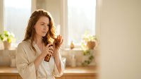 Young Caucasian woman in white bathrobe applying hair serum treatment in bright modern bathroom morning skincare routine wellness self-care lifestyle.