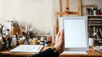 Artist holding tablet with blank screen in creative studio workspace surrounded by art supplies and wooden shelves for design mockups.