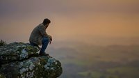 Man sitting on rocky cliff edge overlooking misty valley landscape at sunset, contemplative mood, suitable for mental health, lifestyle, and motivational content.