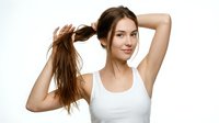 Young woman with long brown hair, fair complexion, wearing white sleeveless top, holding hair in ponytail with both hands raised, smiling at camera against white background.