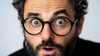 Surprised bearded man with round glasses and wide eyes expressing shock and amazement against light background. Close up portrait showing genuine emotion.