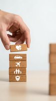 Hand placing wooden block with house icon on stack showing insurance coverage types including auto, travel, family and security protection concepts.