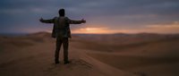 Man in casual jacket standing with arms outstretched on desert sand dune at sunset, expressing freedom and contemplation in vast landscape.