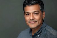 Middle-aged South Asian man with salt and pepper hair wearing gray button-up shirt against dark background, confident smile for professional headshot.
