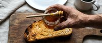 Man's hand drizzling honey onto toasted bread slice on wooden board, breakfast or snack preparation with cup and plate visible.