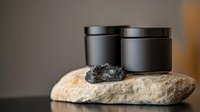 Black cosmetic jars on natural stone with charcoal powder, minimalist spa composition against blurred neutral background for wellness and skincare products.