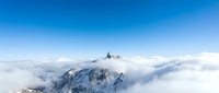 Snow-covered mountain peak emerging through white clouds against clear blue sky creating dramatic alpine landscape for outdoor adventure concepts.