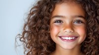 Young girl with curly brown hair and bright smile against light background, radiating joy and confidence. Natural lighting highlights genuine expression.