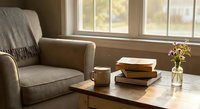 Cozy reading corner with armchair, stacked books and coffee mug by sunny window. Warm natural light creates peaceful atmosphere for relaxation and study.