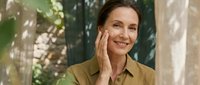 Mature woman applying skincare cream to face outdoors in natural light, anti-aging beauty routine for healthy glowing skin in garden setting.