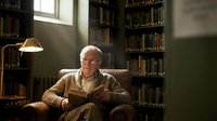 Elderly Caucasian man reading book in cozy library with warm lighting and floor-to-ceiling bookshelves creating peaceful scholarly atmosphere.