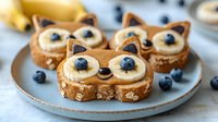 Playful fox-shaped breakfast toast with banana slices, blueberries, and peanut butter on ceramic plate. Creative food art for kids morning meal.