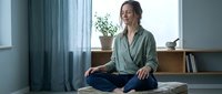 Young woman practicing meditation in peaceful home interior sitting cross-legged on bed wearing casual green shirt with natural lighting and plants.