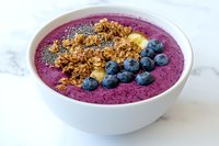Purple smoothie bowl topped with fresh blueberries, crunchy granola and banana slices on white marble background, healthy breakfast concept.