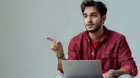 Young South Asian man in burgundy shirt gesturing while speaking, with laptop on lap against neutral background, conveying business conversation or online meeting.