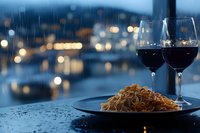 Romantic dinner with pasta and red wine glasses on table with city lights bokeh background during evening rain for date night dining experience.
