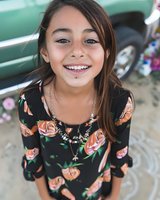 Young Hispanic girl with bright genuine smile wearing floral dress, looking up at camera with natural joy and confidence. Outdoor casual portrait with soft lighting.
