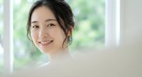 Young Asian woman with natural makeup smiling warmly in bright indoor setting with soft window light. Professional headshot for wellness and lifestyle content.