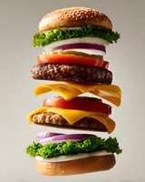 Deconstructed double cheeseburger with beef patties, cheese slices, fresh vegetables and sesame seed buns floating in air against neutral background.