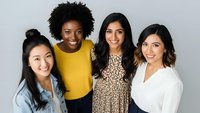 Four diverse young professional women smiling together in casual business attire against neutral background for teamwork and friendship concepts.