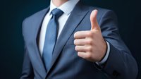 Businessman in navy blue suit showing thumbs up gesture against dark background, conveying approval and positive feedback in professional setting.
