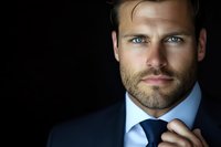 Confident businessman in navy suit adjusting tie against dark background, intense blue eyes and well-groomed beard create professional corporate headshot.