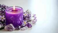Purple candle burning surrounded by amethyst crystals and small white flowers on white background for spa relaxation meditation wellness aromatherapy concept.