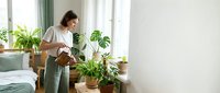 Young woman watering houseplants in bright modern bedroom with green curtains and natural light creating peaceful morning routine atmosphere.