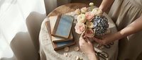 Woman arranging pink roses and eucalyptus bouquet on wooden table with vintage books in soft natural light for romantic home decor and lifestyle photography.