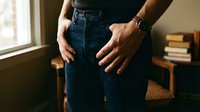 Man in dark denim jeans with hands in pockets, wearing a leather watch, standing indoors in casual style.