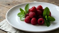 Fresh raspberries with mint leaves arranged on white plate, ideal for food styling, culinary content, healthy eating, dessert recipes, and organic produce marketing.