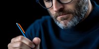 Mature Caucasian man with gray beard and glasses holding pen in dark setting, dramatic lighting highlights thoughtful expression during creative work process.