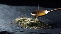 Golden honey drips onto spoon over scattered green hemp protein powder on dark stone surface, creating contrast between liquid and dry ingredients.