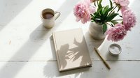 Cozy morning scene with coffee cup, pink peonies in vase, open notebook and pencil on white wooden table with natural sunlight shadows for lifestyle content.