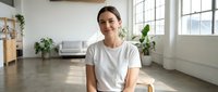 Smiling young Caucasian woman in white t-shirt sitting in bright modern apartment with large windows and plants, natural lighting for lifestyle content.