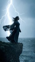 Dramatic woman in flowing dark dress standing on cliff during thunderstorm with lightning strike over stormy ocean waves and heavy rain.