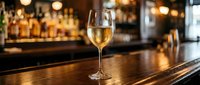 Glass of white wine on wooden bar counter in upscale restaurant with blurred bottles and warm lighting creating elegant dining atmosphere for hospitality industry.