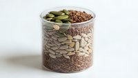 Glass jar filled with layered organic seeds and nuts including pumpkin seeds, flax seeds, and sunflower seeds for healthy cooking and nutrition.
