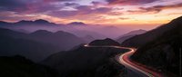 Winding mountain road with light trails at sunset showcasing dramatic purple and pink sky over layered mountain ranges creating scenic landscape view.