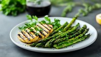 Grilled hasselback potato with fresh green asparagus spears on white plate. Garnished with parsley. Healthy vegetarian dinner or lunch side dish.