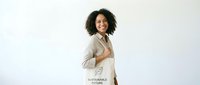Young woman with dark curly hair and medium skin tone wearing beige linen shirt, holding sustainable fabric tote bag, smiling at camera against white background for eco-friendly lifestyle and conscious shopping concepts.