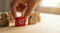 Hand placing a red wooden block with target and arrow icon among human figure blocks, symbolizing leadership, recruitment, and business strategy.