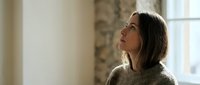 Young woman in cozy sweater looking up thoughtfully by window with natural light streaming in, peaceful moment of reflection and contemplation at home.