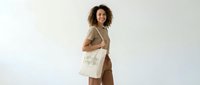Young woman with curly dark hair and medium skin tone wearing beige linen shirt and canvas tote bag, smiling at camera against white background for fashion and lifestyle marketing.