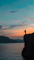 Silhouette of lone fisherman standing on cliff edge against dramatic sunset sky and calm lake waters, mountains in background creating serene outdoor scene.