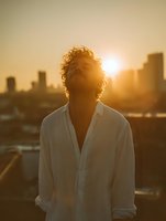 Young man with curly hair in white shirt standing against urban sunset skyline, eyes closed in peaceful moment, golden hour lighting creates atmospheric silhouette.