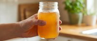 Unrecognizable person holding glass jar filled with golden kombucha or fermented tea beverage in bright kitchen setting with blurred background.