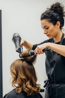 Professional female hairstylist with curly hair styling blonde client with blow dryer and round brush creating voluminous waves in modern salon.