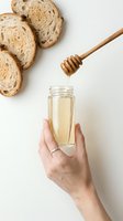 Honey dripping from wooden dipper into glass jar held by woman's hand, with sliced bread on white background. Food styling for cooking and baking.