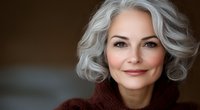 Confident mature woman with silver gray wavy hair and natural makeup wearing brown knit sweater smiling warmly against dark background, beauty and aging gracefully concept.